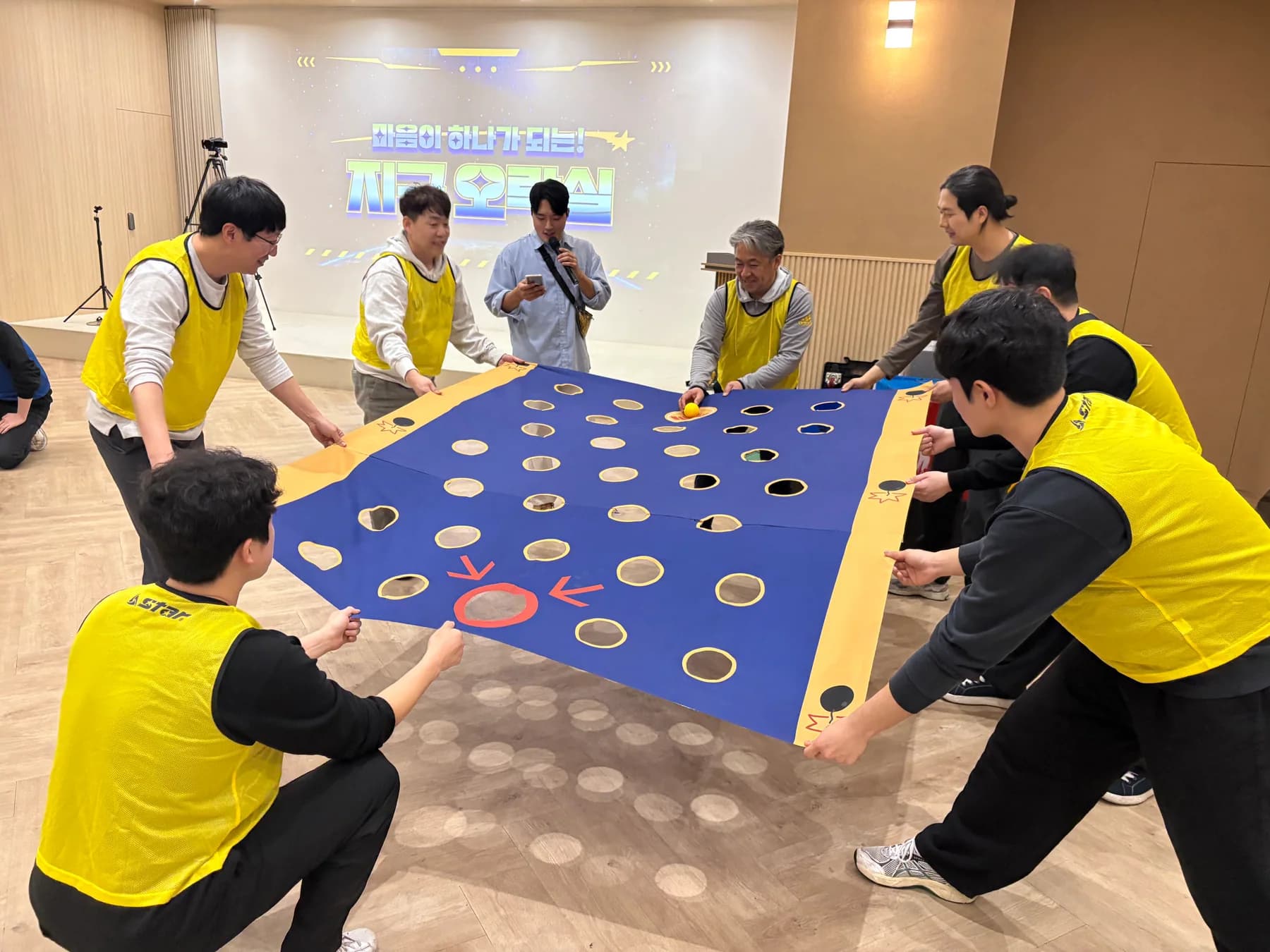 노란 팀 참가자들이 구멍이 뚫린 대형 천을 함께 잡고 공을 골대까지 이동하는 실제 팀빌딩 현장