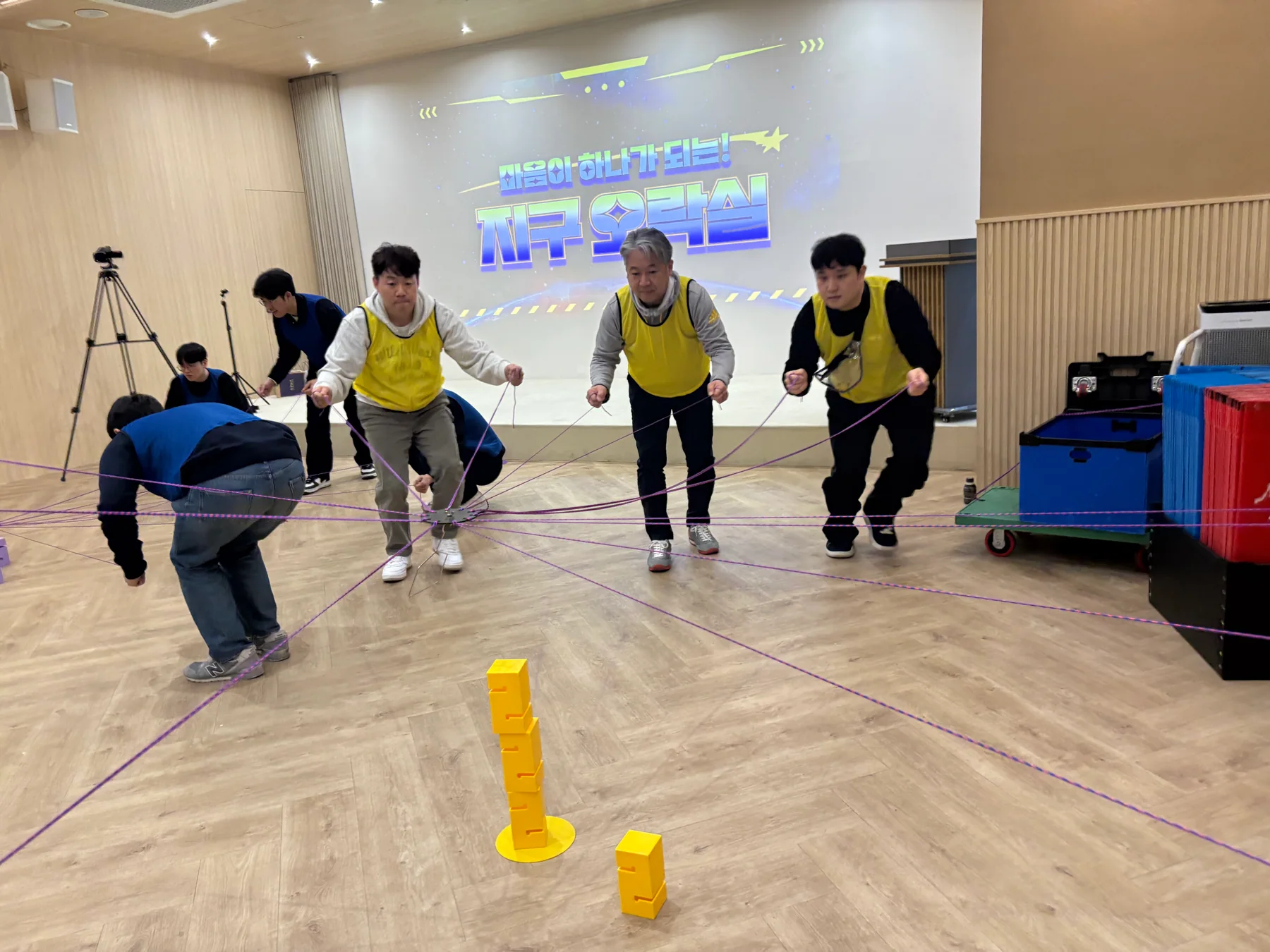 노란 팀 조끼를 입은 참가자들이 줄을 함께 조작해 블록 탑을 쌓는 실제 팀빌딩 현장